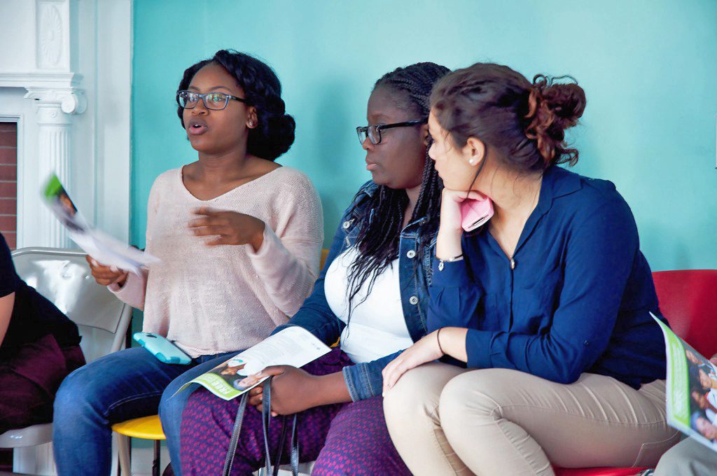 Three young people sitting together in a room with blue walls, one of them speaking and holding a piece of paper