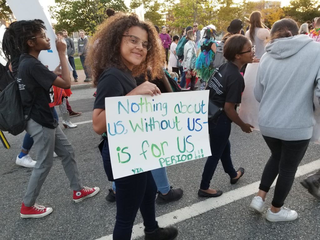 A young person at a march holding a handmade sign that reads "Nothing about us, without us – is for us. PERIOD!!!"