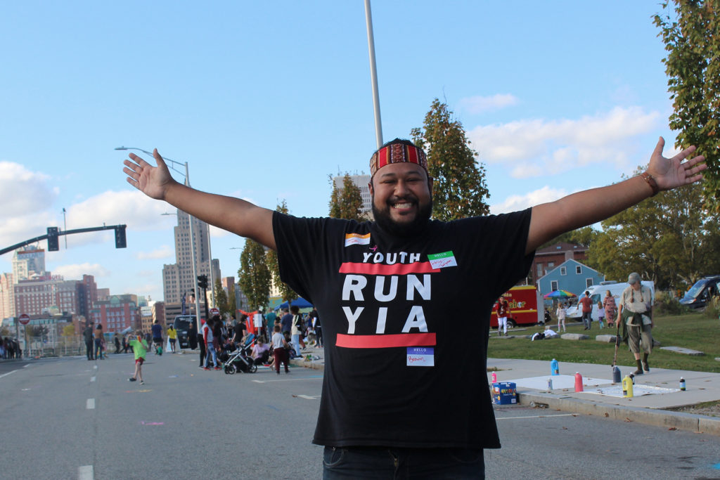 Elliot standing in the street at an event on a sunny day smiling big with his arms outstretched